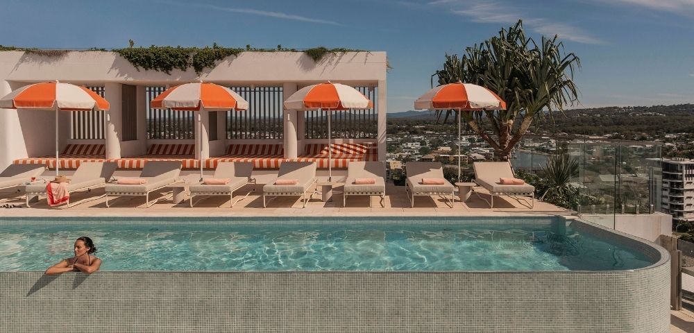 A person rests in a turquoise swimming pool with a tiled edge, overlooking a sunny rooftop lounge area with beige lounge chairs and orange and white striped umbrellas.