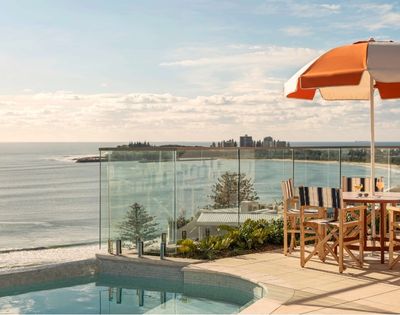Outdoor seating area with a table, chairs, and an orange and white striped umbrella, overlooking a calm ocean with a distant coastline and buildings.
