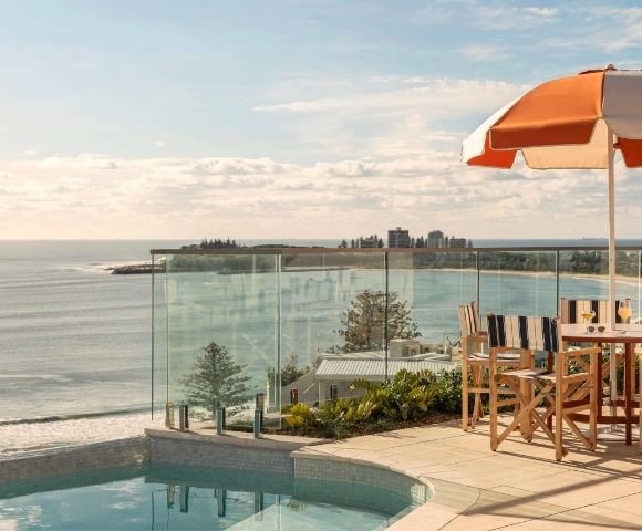 Outdoor seating area with a table, chairs, and an orange and white striped umbrella, overlooking a calm ocean with a distant coastline and buildings.