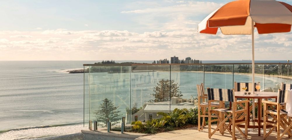 Outdoor seating area with a table, chairs, and an orange and white striped umbrella, overlooking a calm ocean with a distant coastline and buildings.