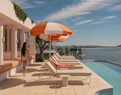 A woman in a light-colored swimsuit walks on a rooftop patio beside a row of lounge chairs and orange and white umbrellas, with an infinity pool and a coastal cityscape in the background under a blue sky.