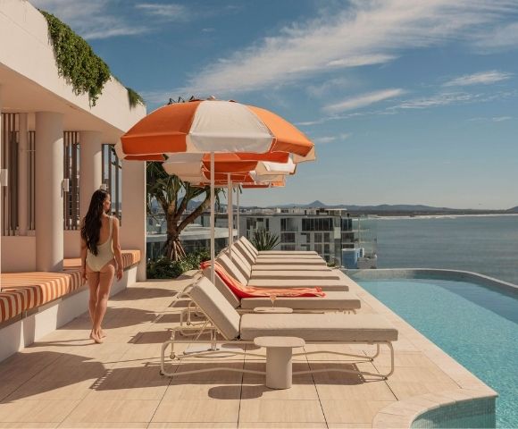 A woman in a light-colored swimsuit walks on a rooftop patio beside a row of lounge chairs and orange and white umbrellas, with an infinity pool and a coastal cityscape in the background under a blue sky.