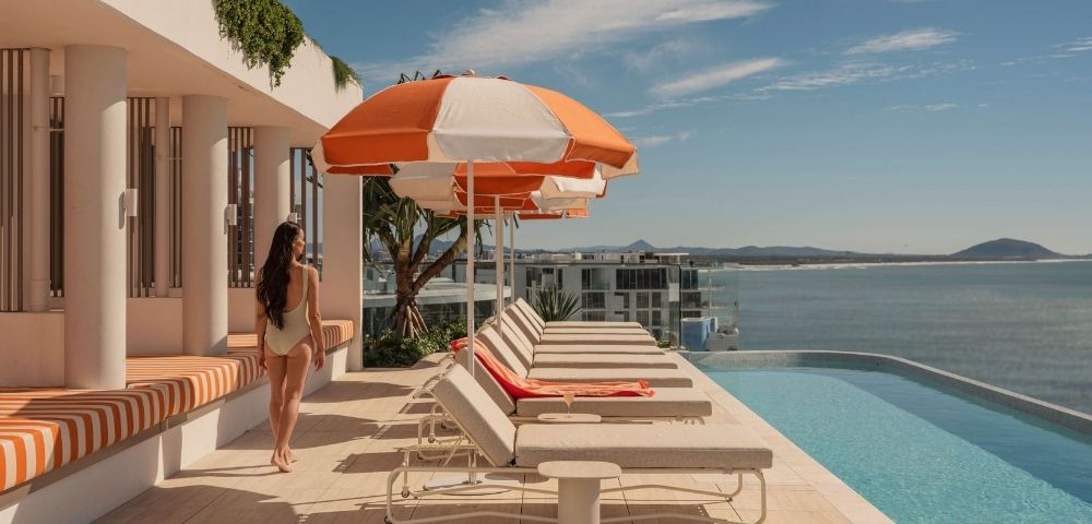 A woman in a light-colored swimsuit walks on a rooftop patio beside a row of lounge chairs and orange and white umbrellas, with an infinity pool and a coastal cityscape in the background under a blue sky.