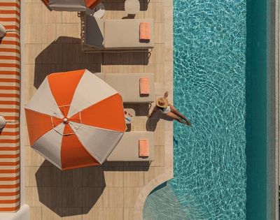 Aerial view of a person in a sun hat sitting on the edge of a bright blue swimming pool, with their legs dangling in the water, next to beige lounge chairs with orange towels and a large orange and white umbrella.