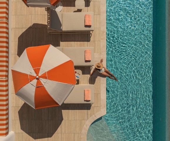 Aerial view of a person in a sun hat sitting on the edge of a bright blue swimming pool, with their legs dangling in the water, next to beige lounge chairs with orange towels and a large orange and white umbrella.