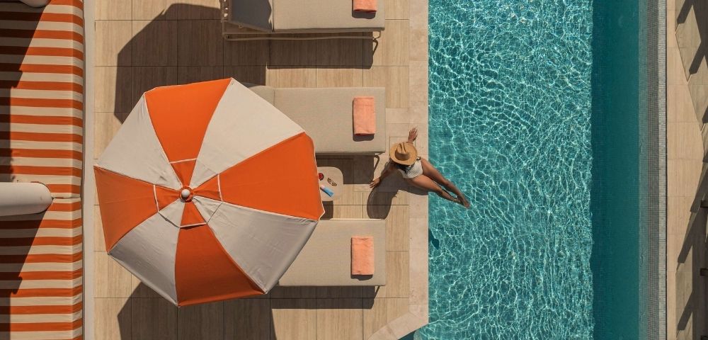 Aerial view of a person in a sun hat sitting on the edge of a bright blue swimming pool, with their legs dangling in the water, next to beige lounge chairs with orange towels and a large orange and white umbrella.