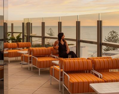 A woman in a dark dress sits on a striped orange outdoor lounge chair, holding a pink drink and looking out at the ocean during sunset.