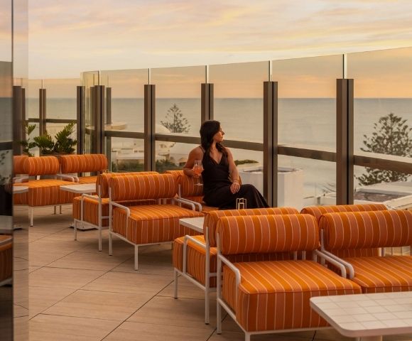 A woman in a dark dress sits on a striped orange outdoor lounge chair, holding a pink drink and looking out at the ocean during sunset.