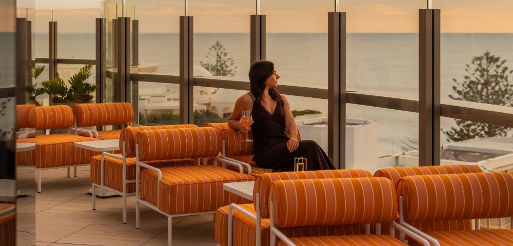 A woman in a dark dress sits on a striped orange outdoor lounge chair, holding a pink drink and looking out at the ocean during sunset.