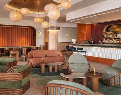 Modern lounge area with curved orange sofa, green upholstered chairs with rattan frames, marble-topped tables, and a long bar with shelves of liquor bottles.
