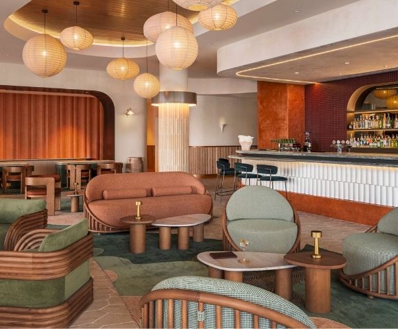 Modern lounge area with curved orange sofa, green upholstered chairs with rattan frames, marble-topped tables, and a long bar with shelves of liquor bottles.