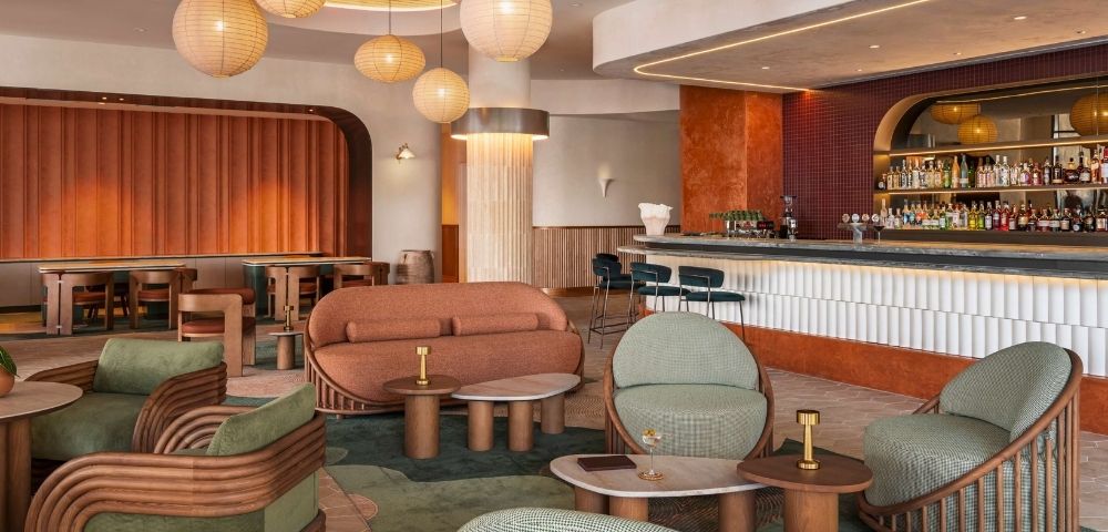 Modern lounge area with curved orange sofa, green upholstered chairs with rattan frames, marble-topped tables, and a long bar with shelves of liquor bottles.