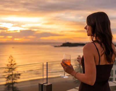 Woman with dark hair holding a glass of rosé wine on a balcony, looking out at the ocean during a vibrant sunset.