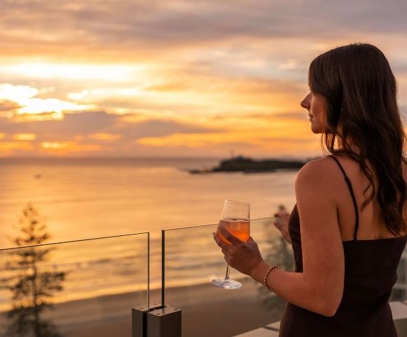 Woman with dark hair holding a glass of rosé wine on a balcony, looking out at the ocean during a vibrant sunset.