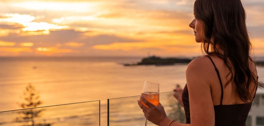 Woman with dark hair holding a glass of rosé wine on a balcony, looking out at the ocean during a vibrant sunset.