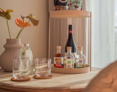 A decorative vase with orange anthurium flowers stands next to a bottle of water and two glasses, with a tray holding small bottles of liquor and a larger bottle of wine in the background.