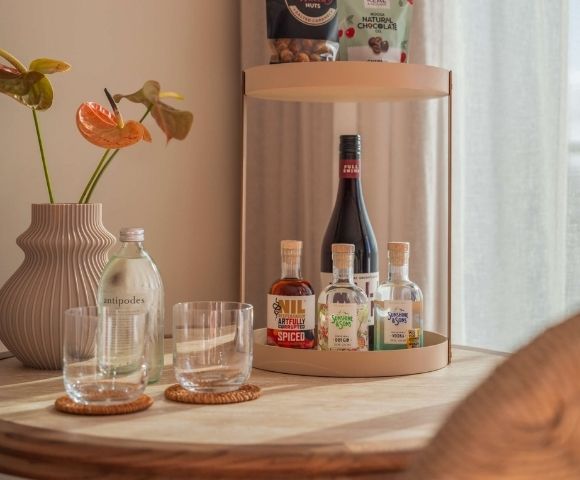 A decorative vase with orange anthurium flowers stands next to a bottle of water and two glasses, with a tray holding small bottles of liquor and a larger bottle of wine in the background.