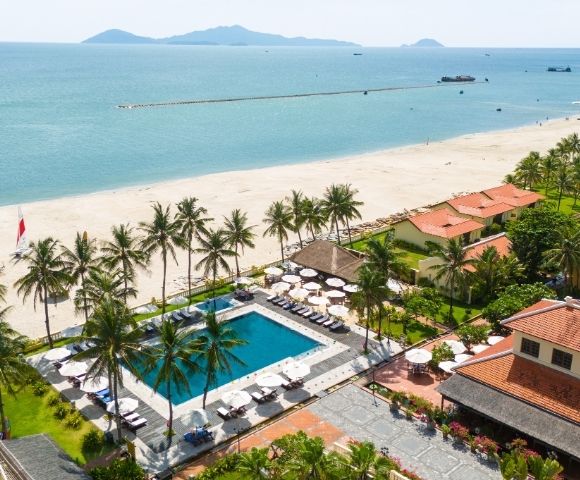Aerial view of a tropical resort with a large swimming pool and lounge chairs, surrounded by palm trees and low-rise buildings with terracotta roofs, leading to a white sandy beach and the turquoise ocean under a clear sky.
