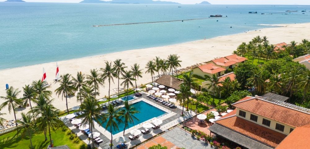 Aerial view of a tropical resort with a large swimming pool and lounge chairs, surrounded by palm trees and low-rise buildings with terracotta roofs, leading to a white sandy beach and the turquoise ocean under a clear sky.