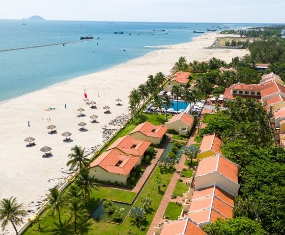 Aerial view of a tropical resort with terracotta-roofed buildings nestled among palm trees, a swimming pool, and a pristine white sand beach bordering a calm turquoise ocean dotted with small boats and a distant breakwater.