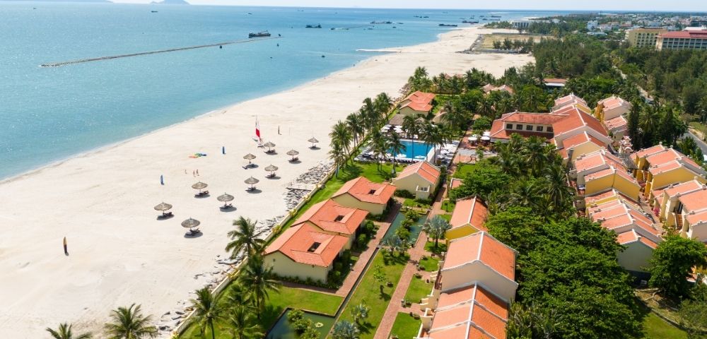 Aerial view of a tropical resort with terracotta-roofed buildings nestled among palm trees, a swimming pool, and a pristine white sand beach bordering a calm turquoise ocean dotted with small boats and a distant breakwater.