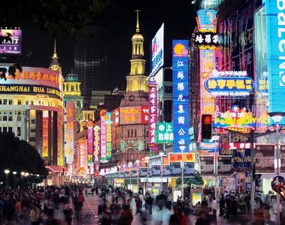 A bustling street scene at night, with a crowd of people walking along a sidewalk lined with buildings covered in bright, colorful neon signs.