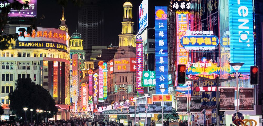 A bustling street scene at night, with a crowd of people walking along a sidewalk lined with buildings covered in bright, colorful neon signs.