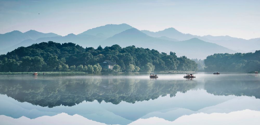Three small boats navigate a calm lake with reflections of lush green trees and layered blue mountains under a clear, hazy sky.