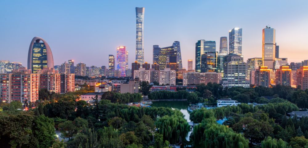 Modern city skyline at dusk with lush green trees and a winding body of water in the foreground.