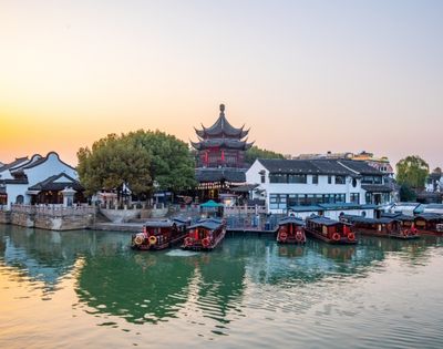Traditional boats docked along a canal in front of white buildings and a multi-tiered, dark-roofed pagoda at sunset.