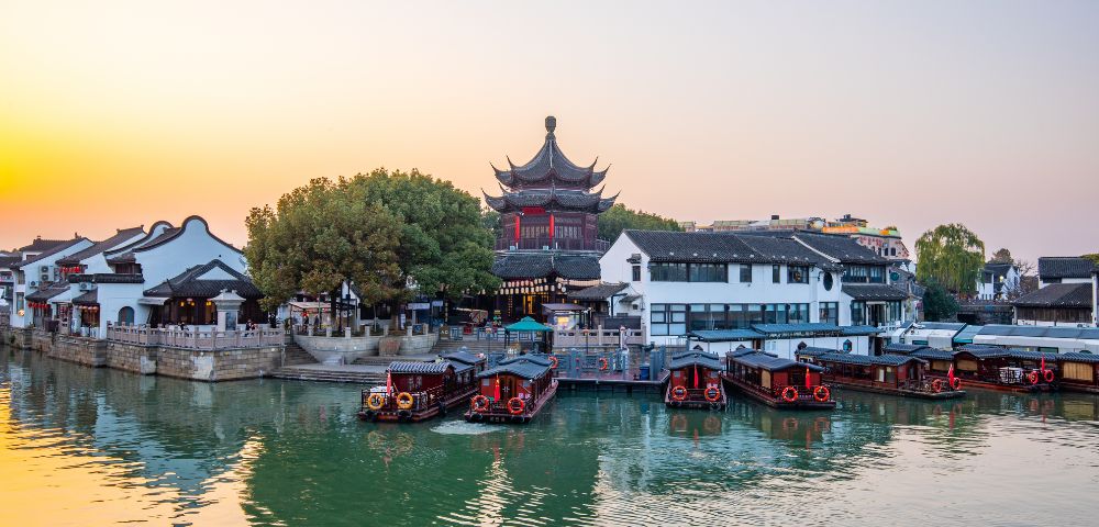 Traditional boats docked along a canal in front of white buildings and a multi-tiered, dark-roofed pagoda at sunset.