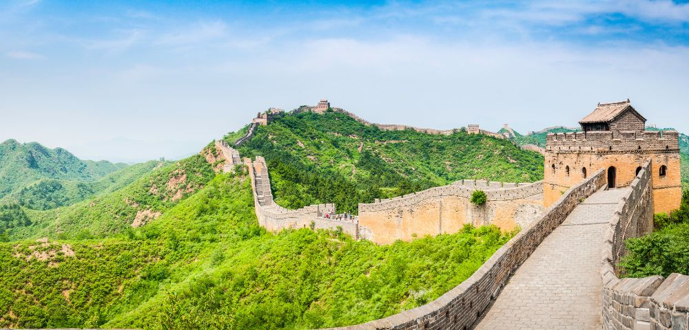 The Great Wall of China winds along lush green, tree-covered mountains under a clear blue sky.
