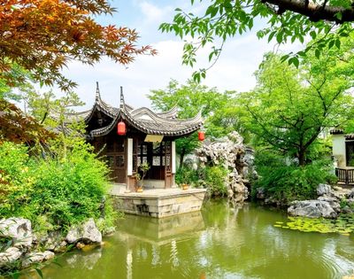 A traditional Chinese pavilion with a curved roof sits on the edge of a pond in a lush garden.