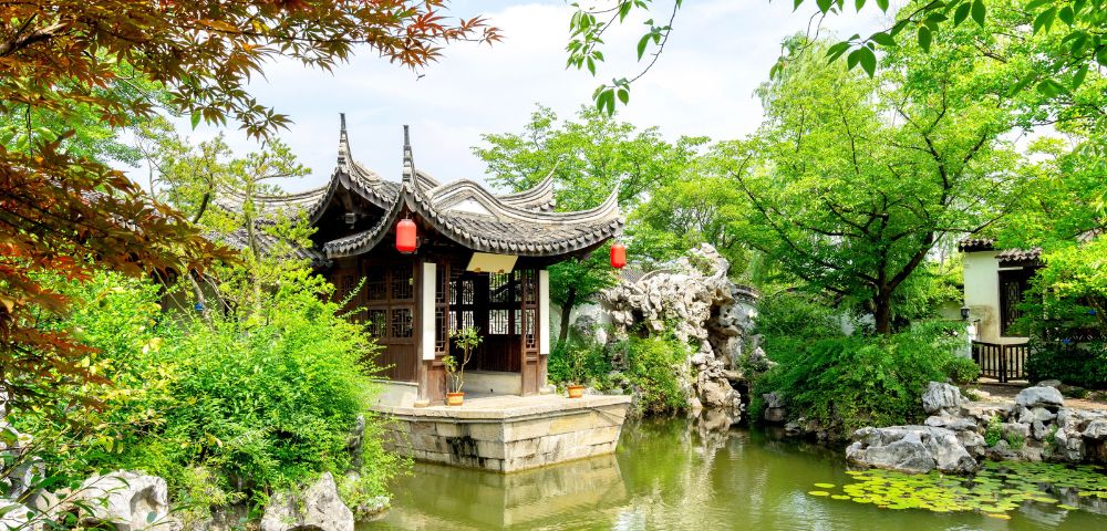 A traditional Chinese pavilion with a curved roof sits on the edge of a pond in a lush garden.