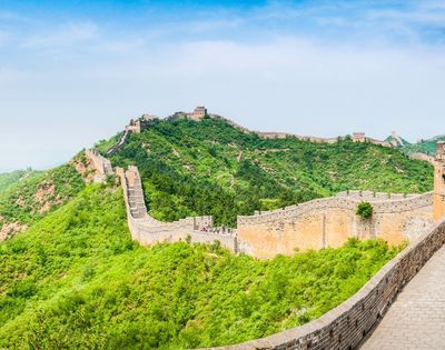 The Great Wall of China winds along lush green, tree-covered mountains under a clear blue sky.