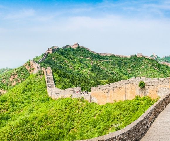 The Great Wall of China winds along lush green, tree-covered mountains under a clear blue sky.