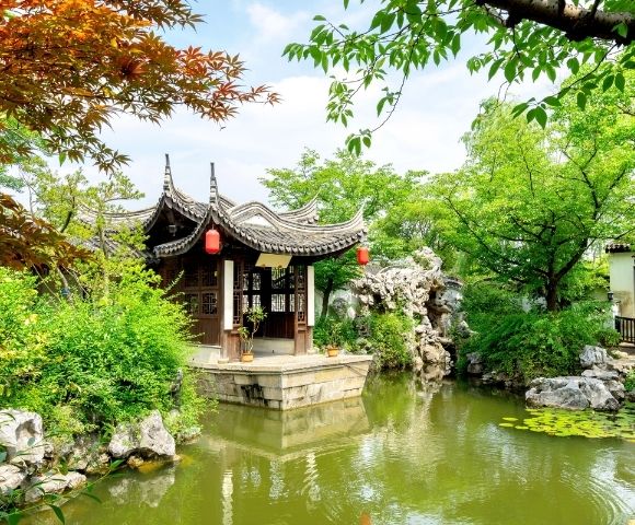 A traditional Chinese pavilion with a curved roof sits on the edge of a pond in a lush garden.