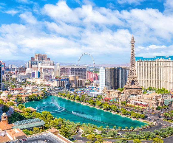 Panoramic view of the Las Vegas Strip featuring a replica Eiffel Tower, a large hotel with a blue roof, a Ferris wheel, and a vibrant blue fountain display in a central canal, set against a bright blue sky with scattered clouds.