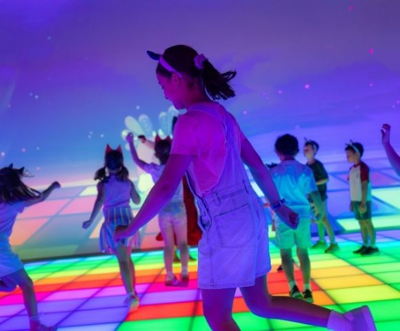 Children dance on a brightly lit, interactive floor that displays a colorful grid pattern.