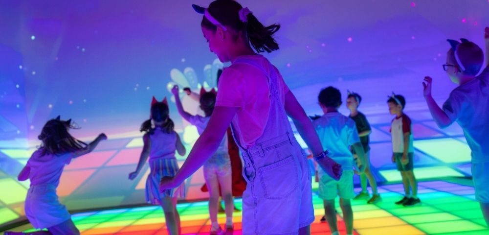 Children dance on a brightly lit, interactive floor that displays a colorful grid pattern.
