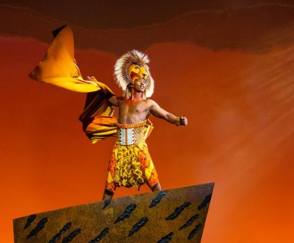 A performer in a lion costume with a flowing yellow cape stands on a rock formation against an orange sky.