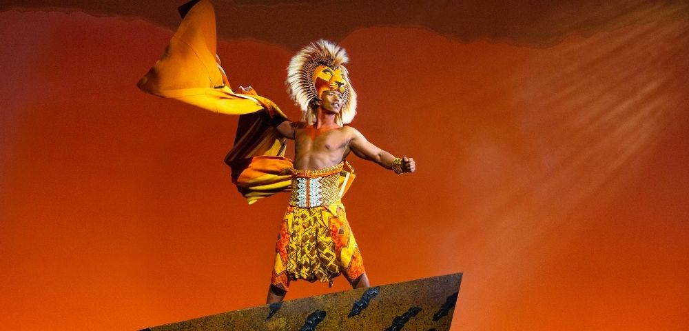 A performer in a lion costume with a flowing yellow cape stands on a rock formation against an orange sky.