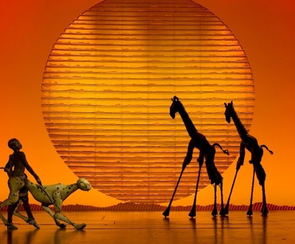 Silhouetted giraffes stand on a stage in front of a large, textured, orange sun, while a costumed figure leads a spotted puppet creature.