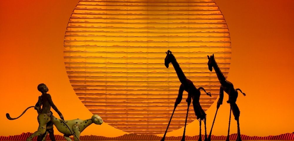 Silhouetted giraffes stand on a stage in front of a large, textured, orange sun, while a costumed figure leads a spotted puppet creature.