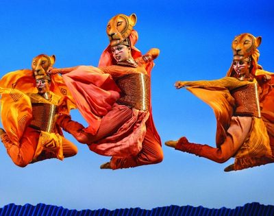 Performers in elaborate lion costumes with flowing orange and red fabrics dance in mid-air against a bright blue sky, suggesting a theatrical production.