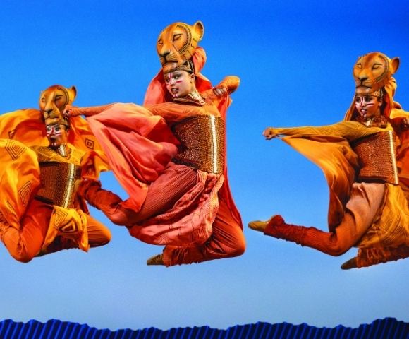 Performers in elaborate lion costumes with flowing orange and red fabrics dance in mid-air against a bright blue sky, suggesting a theatrical production.