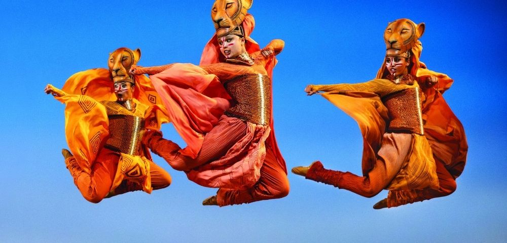 Performers in elaborate lion costumes with flowing orange and red fabrics dance in mid-air against a bright blue sky, suggesting a theatrical production.