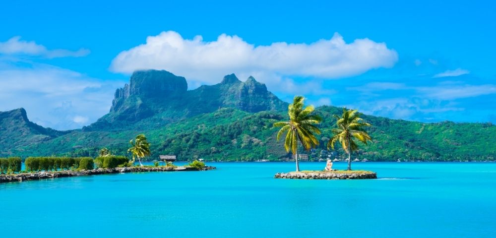 Bright tropical lagoon with turquoise water, palm trees on tiny islets, and lush volcanic mountains beneath a serene, cloud-dotted blue sky.
