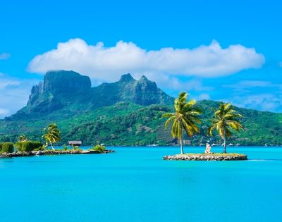 Bright tropical lagoon with turquoise water, palm trees on tiny islets, and lush volcanic mountains beneath a serene, cloud-dotted blue sky.