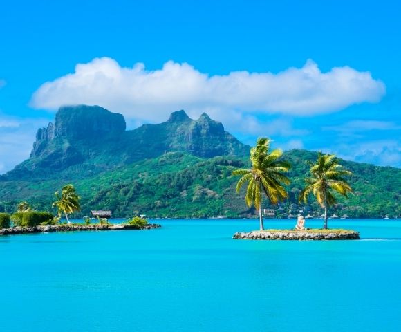 Bright tropical lagoon with turquoise water, palm trees on tiny islets, and lush volcanic mountains beneath a serene, cloud-dotted blue sky.